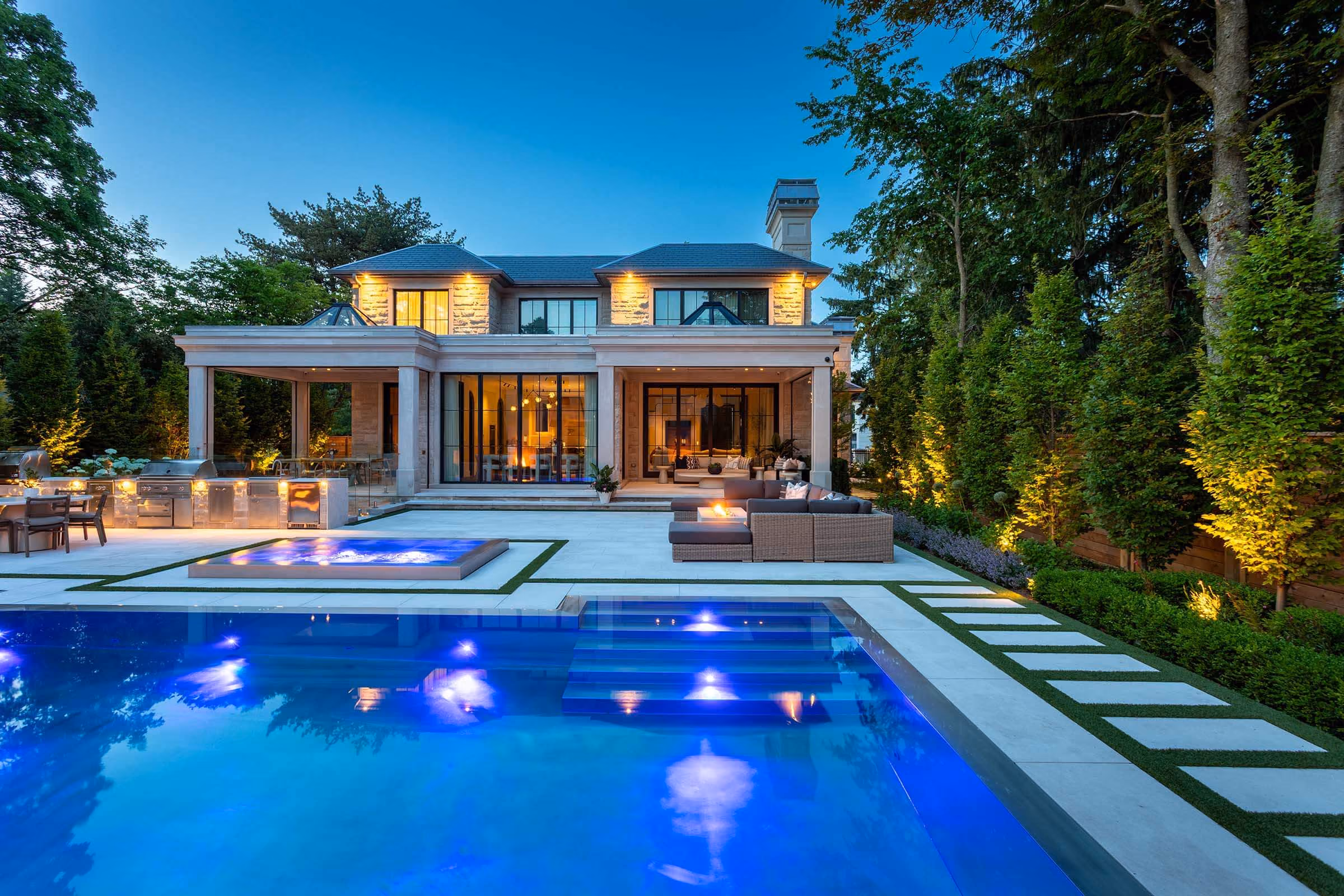 Outdoor pool and hot tub lit purple at twilight in front of a pergola with fire features and seating.