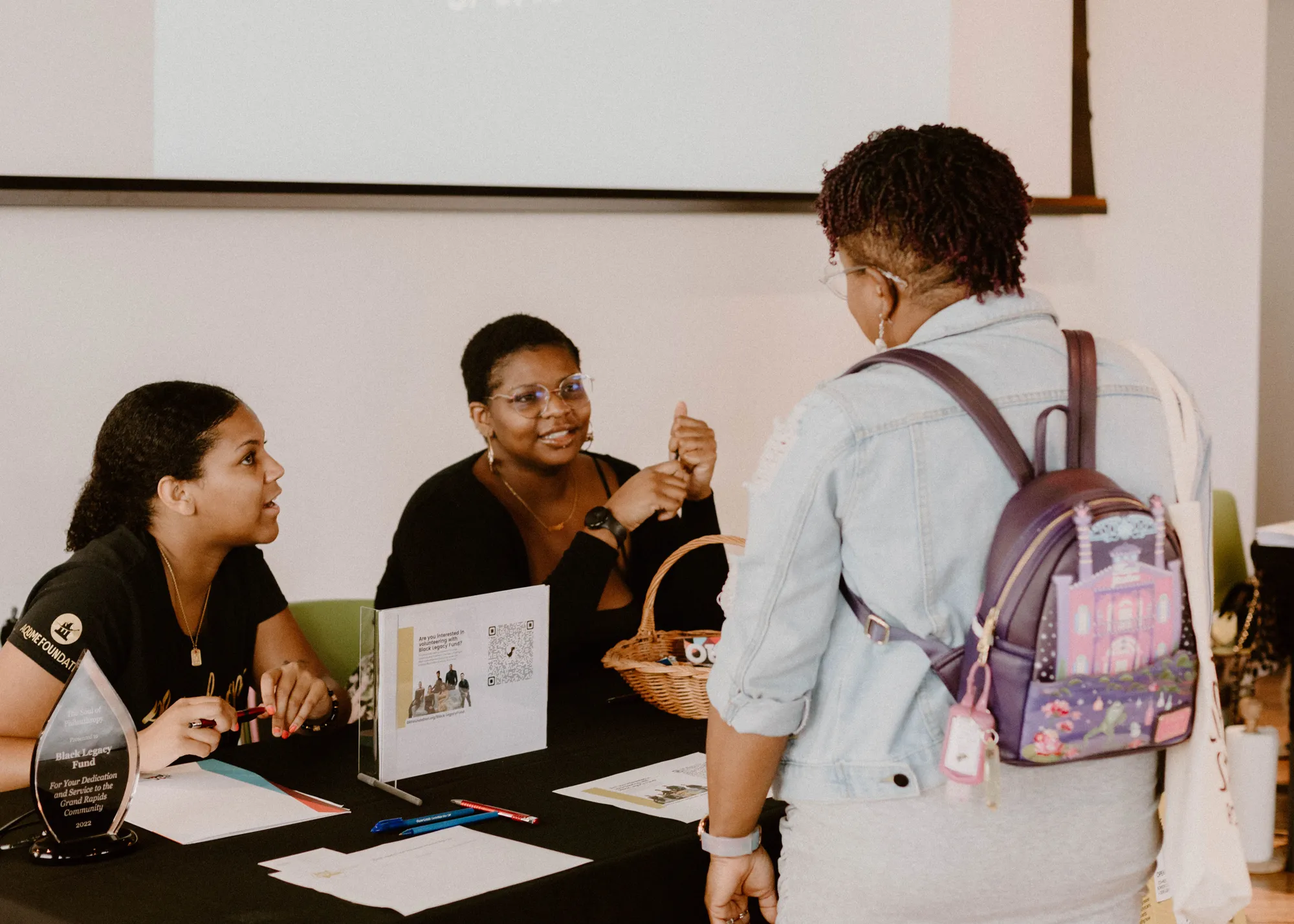 Two women sit behind a table talking to another woman standing with a backpack in front of them.