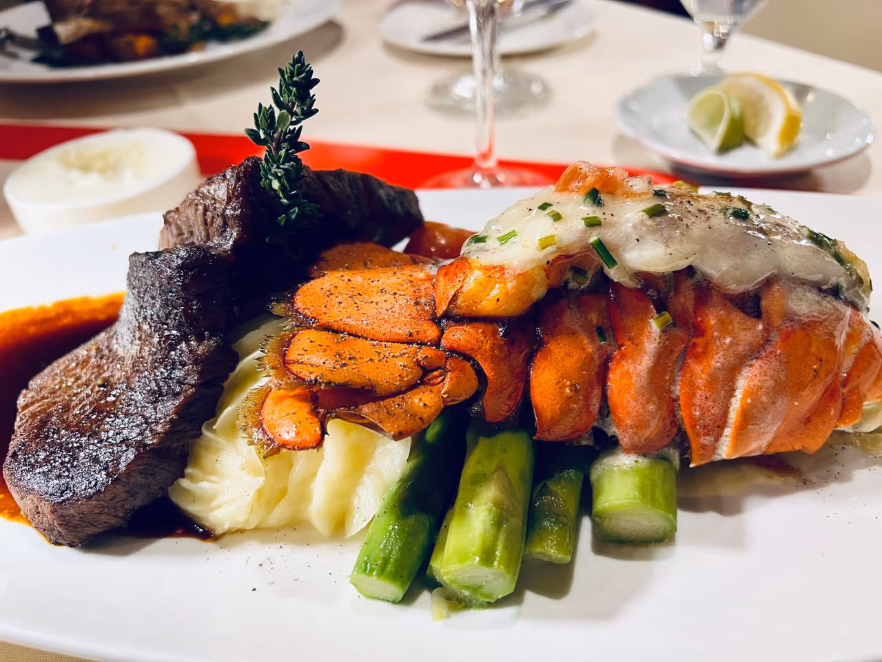 Plated surf and turf with grilled lobster tail, steak, mashed potatoes, and asparagus on a white plate.