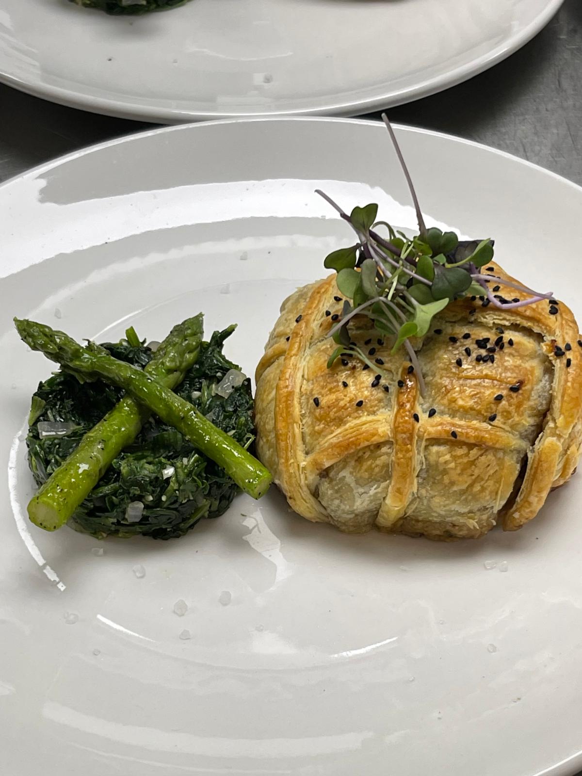 Golden puff pastry with microgreens, served with sautéed spinach and two asparagus spears on white plate.