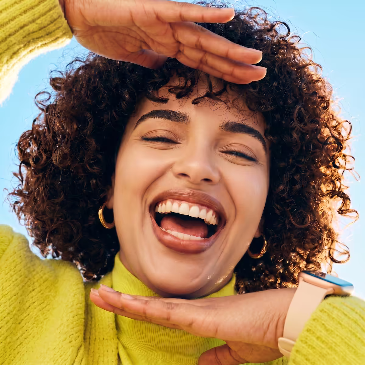 Smiling woman with curly hair framing her face with her hands wearing a yellow sweater and watch.
