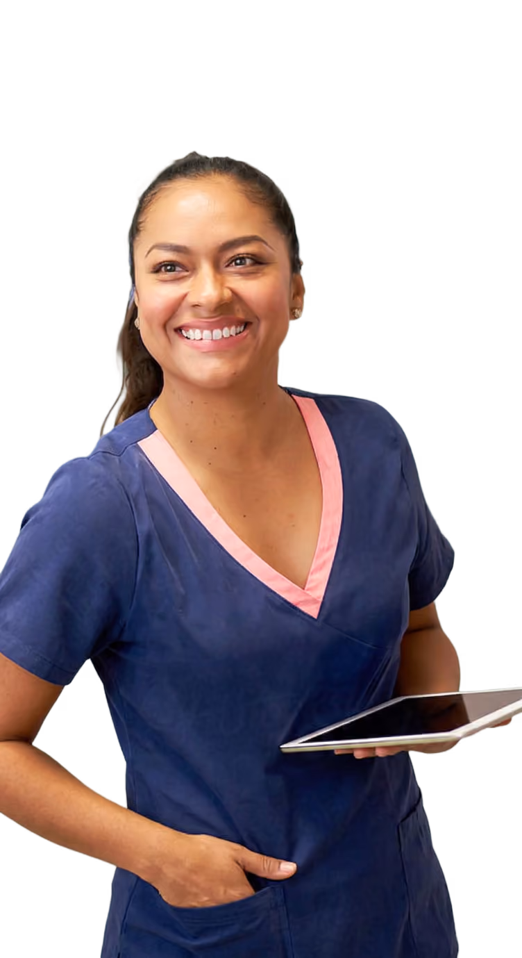 Smiling healthcare worker in blue scrubs holding a tablet with one hand in pocket.
