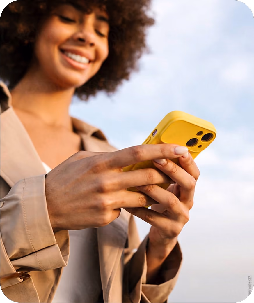 Smiling woman holding and using a yellow smartphone outdoors with a sky background.