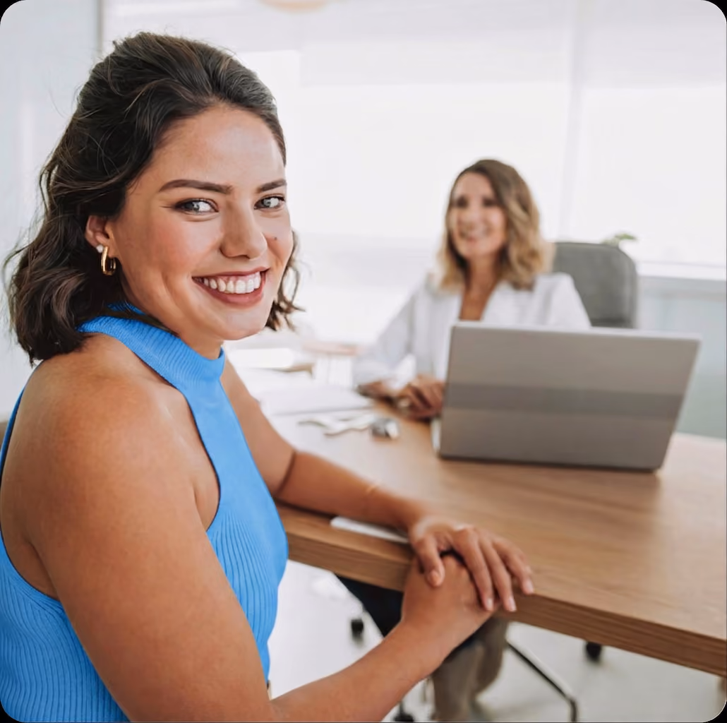 Smiling woman in blue sleeveless top sitting at desk with another woman and laptop in background.