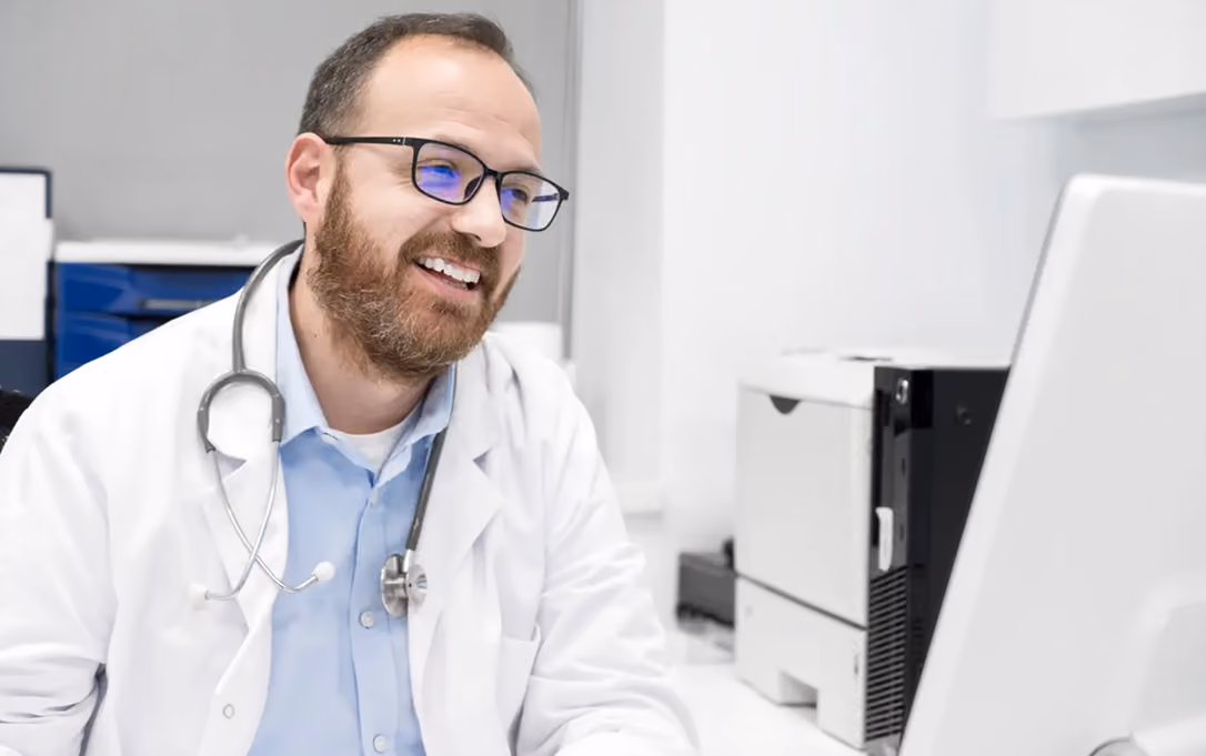 Smiling male doctor with glasses and stethoscope looking at computer screen in clinic office.