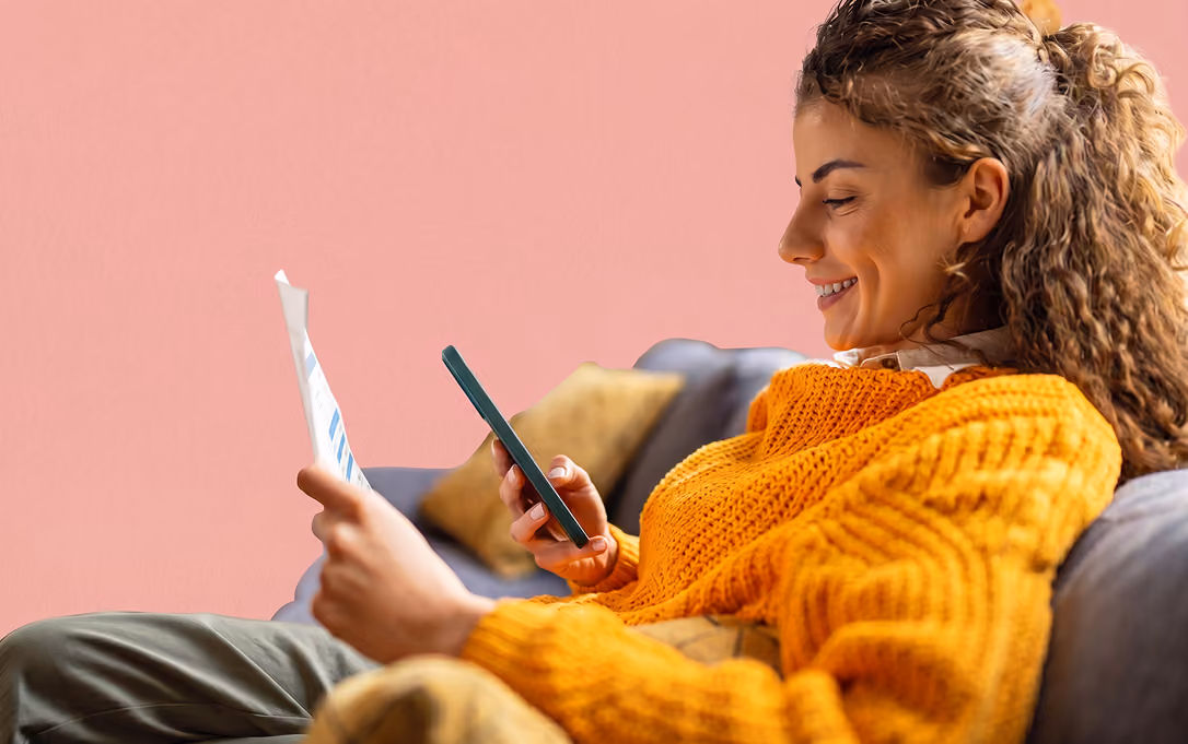 Smiling woman in orange sweater sitting on couch, holding paper in one hand and phone in the other.