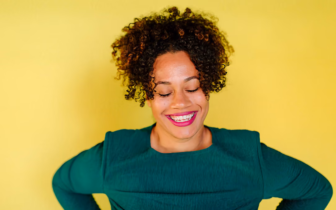 Smiling woman with curly hair and pink lipstick wearing a green top against yellow background.