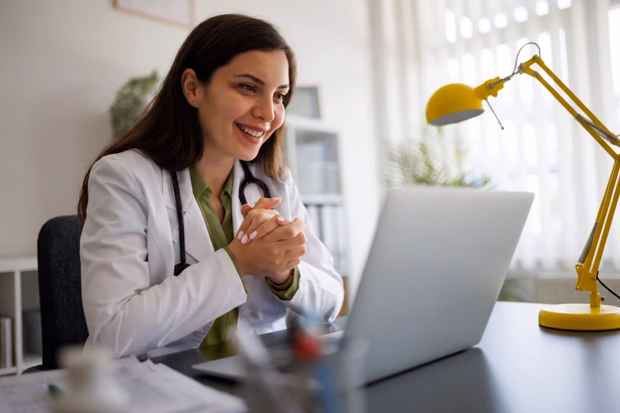 Female doctor in white coat smiling and using a laptop at a desk with a yellow lamp.