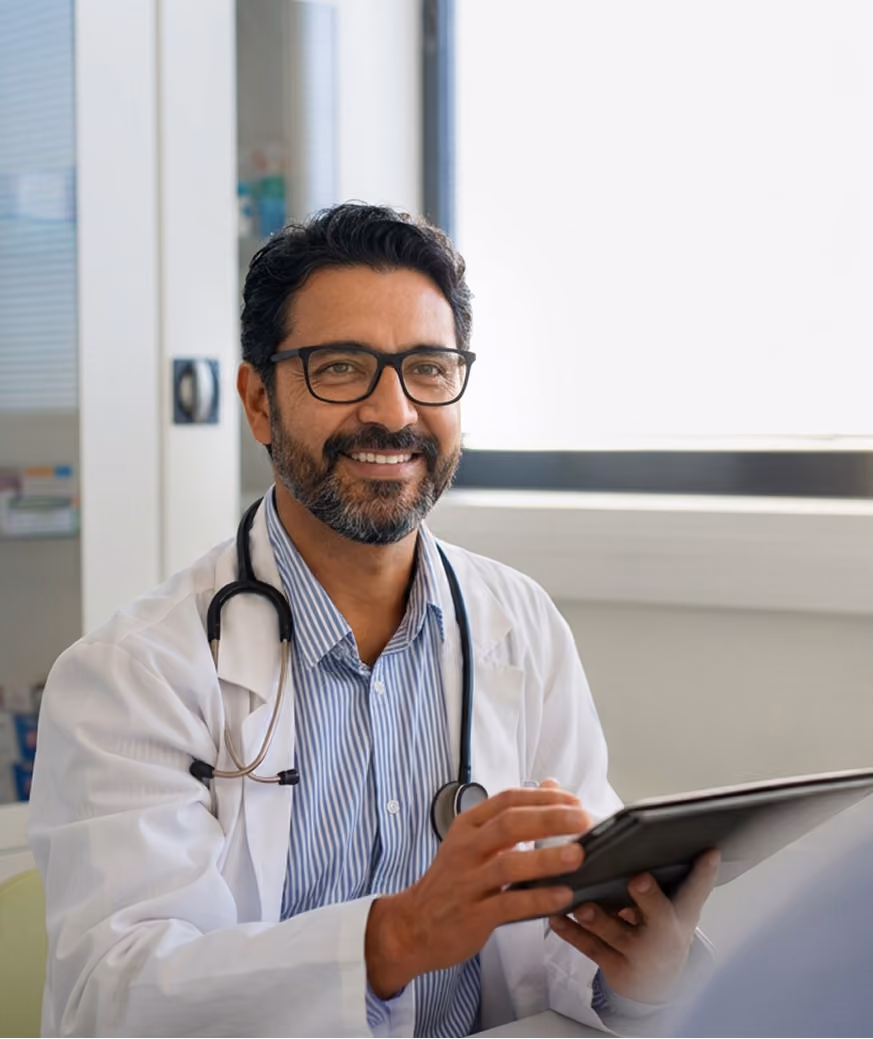 Smiling male doctor with glasses and stethoscope using a tablet in a bright office.