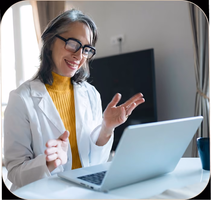 Smiling woman wearing glasses and white coat using a laptop during a video call at home.