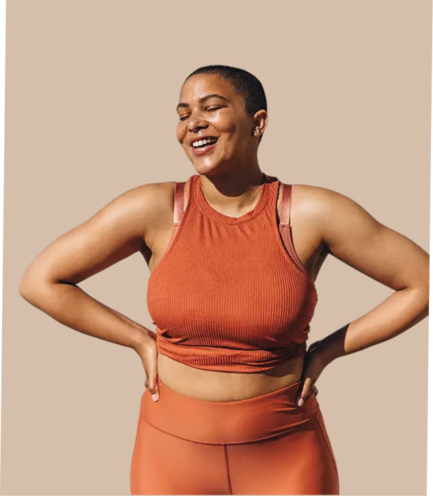 Smiling woman in orange workout top and leggings with hands on hips against beige background.