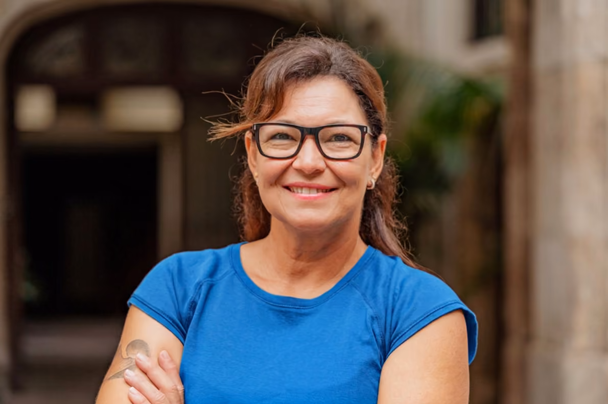 Smiling woman wearing glasses and blue shirt with arms crossed in an outdoor setting.