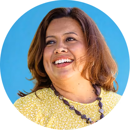 Smiling woman with shoulder-length hair wearing a yellow top and a beaded necklace against a blue background.