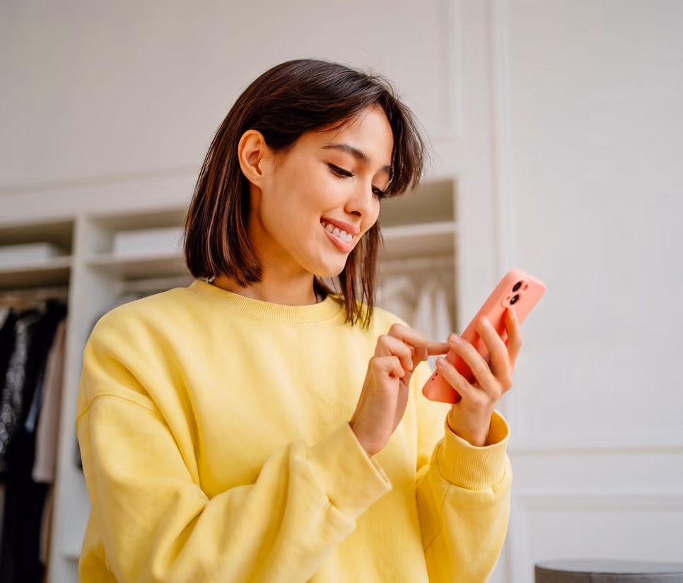 Smiling woman in yellow sweater uses a pink smartphone indoors near a wardrobe.