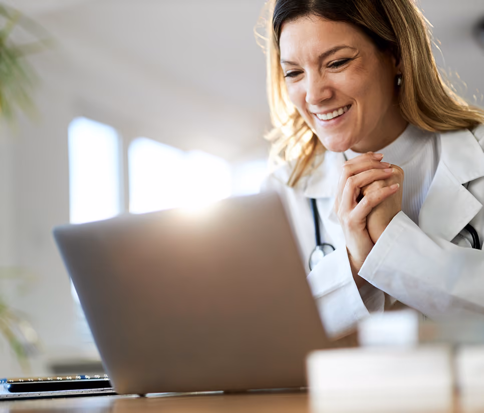 Smiling female doctor in white coat looking at laptop screen during a video call.