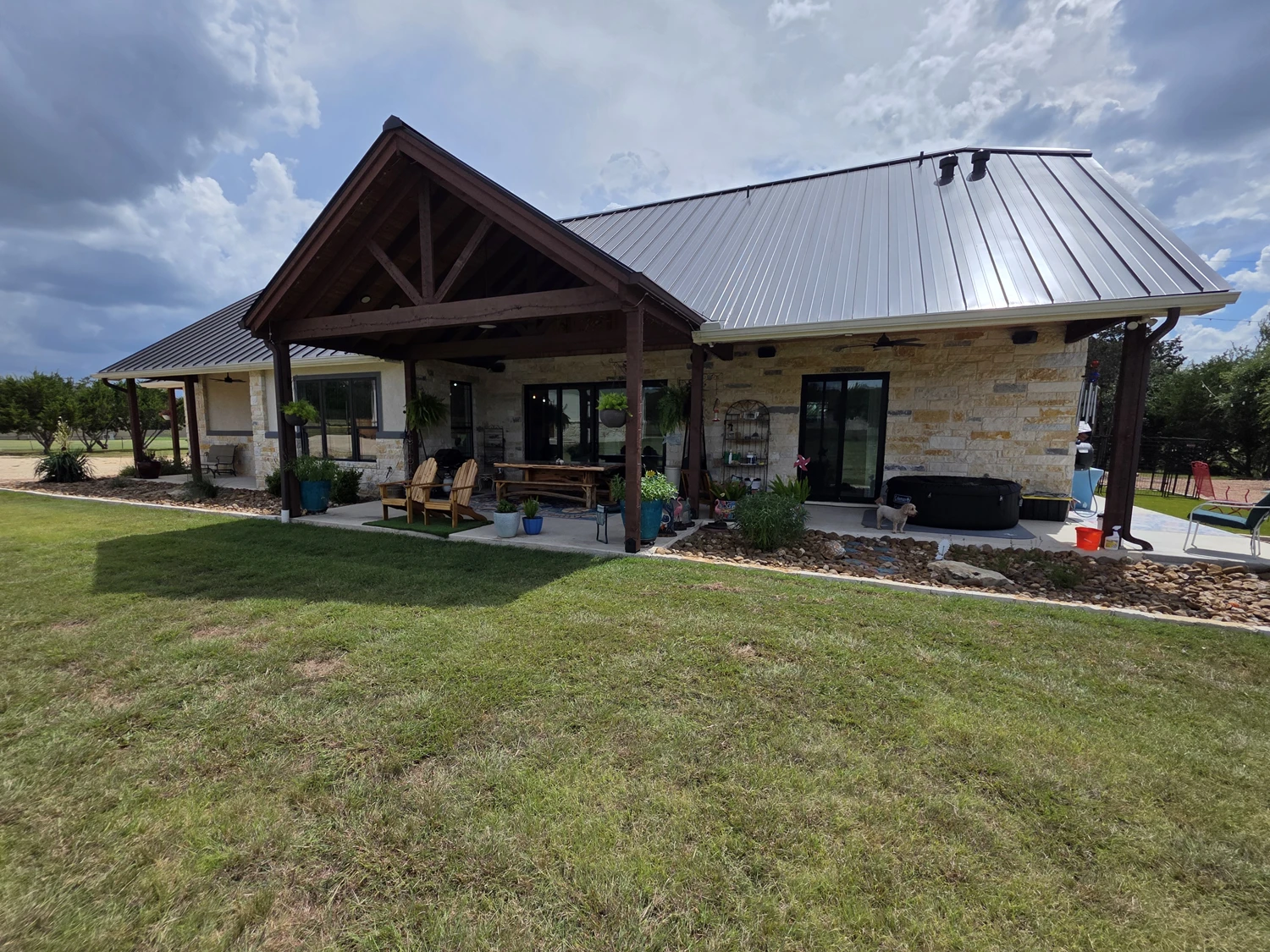 Covered rear patio with a timber gable, outdoor kitchen, and hot tub behind the custom home