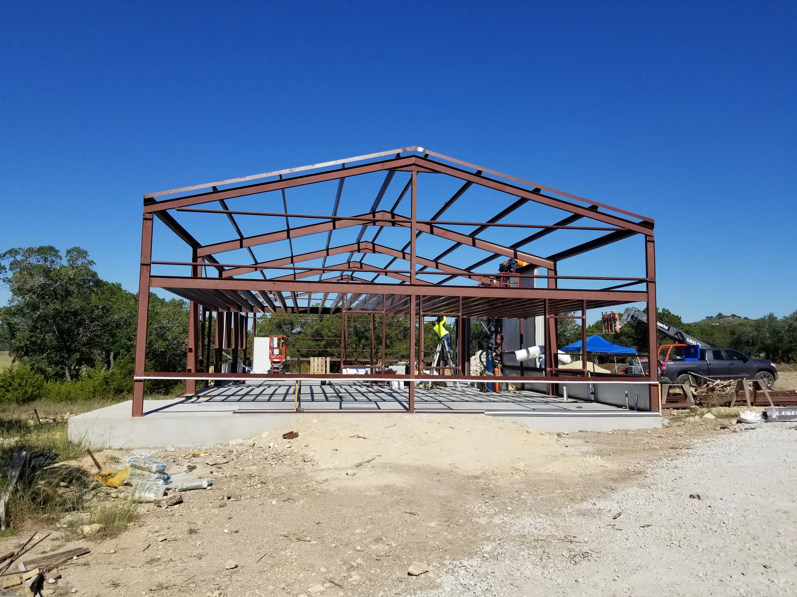 Gable end of the steel frame standing over the finished slab with the crew and a scissor lift on site