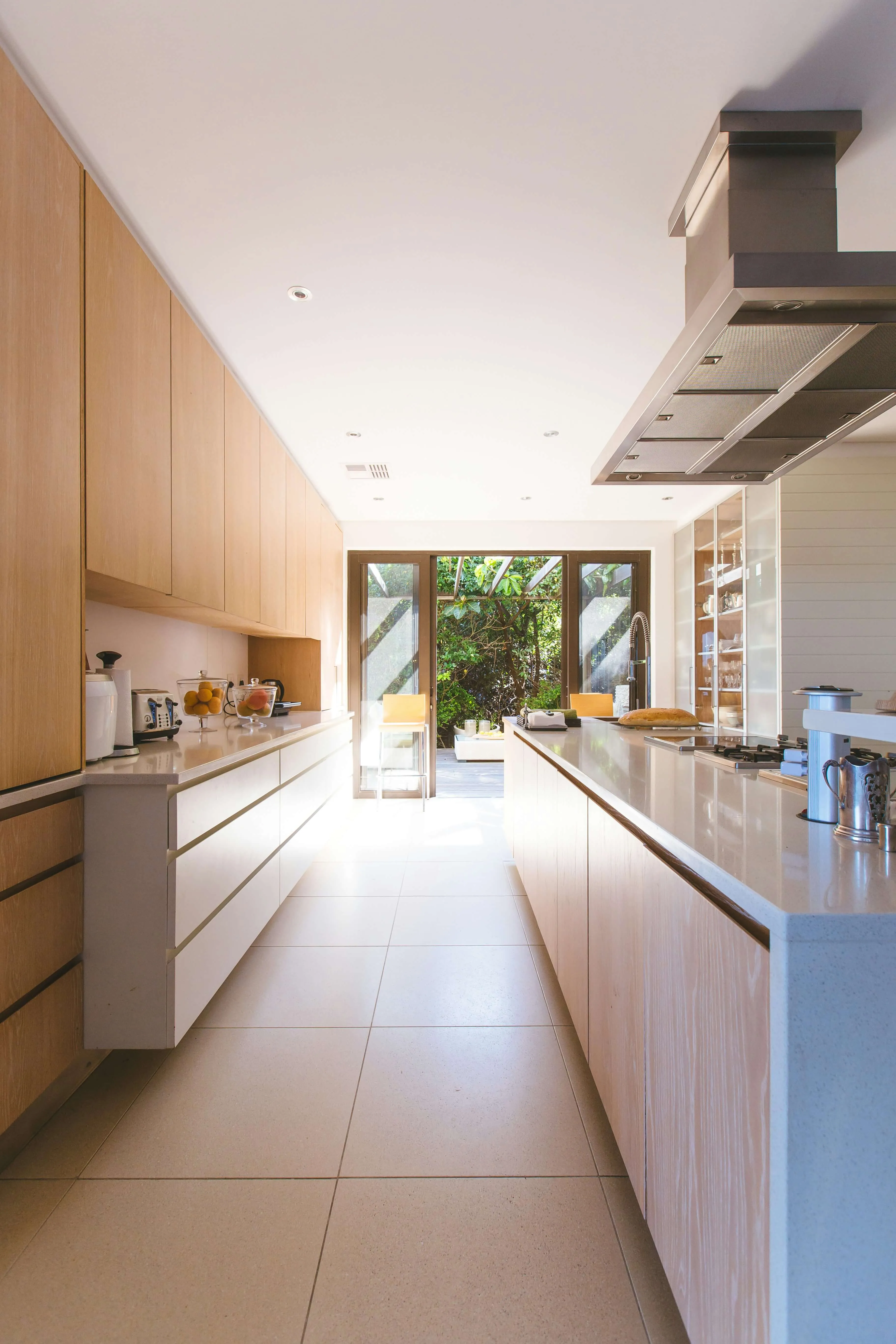 Bright modern kitchen with light wood cabinets, white counters, and a view of a green outdoor patio.