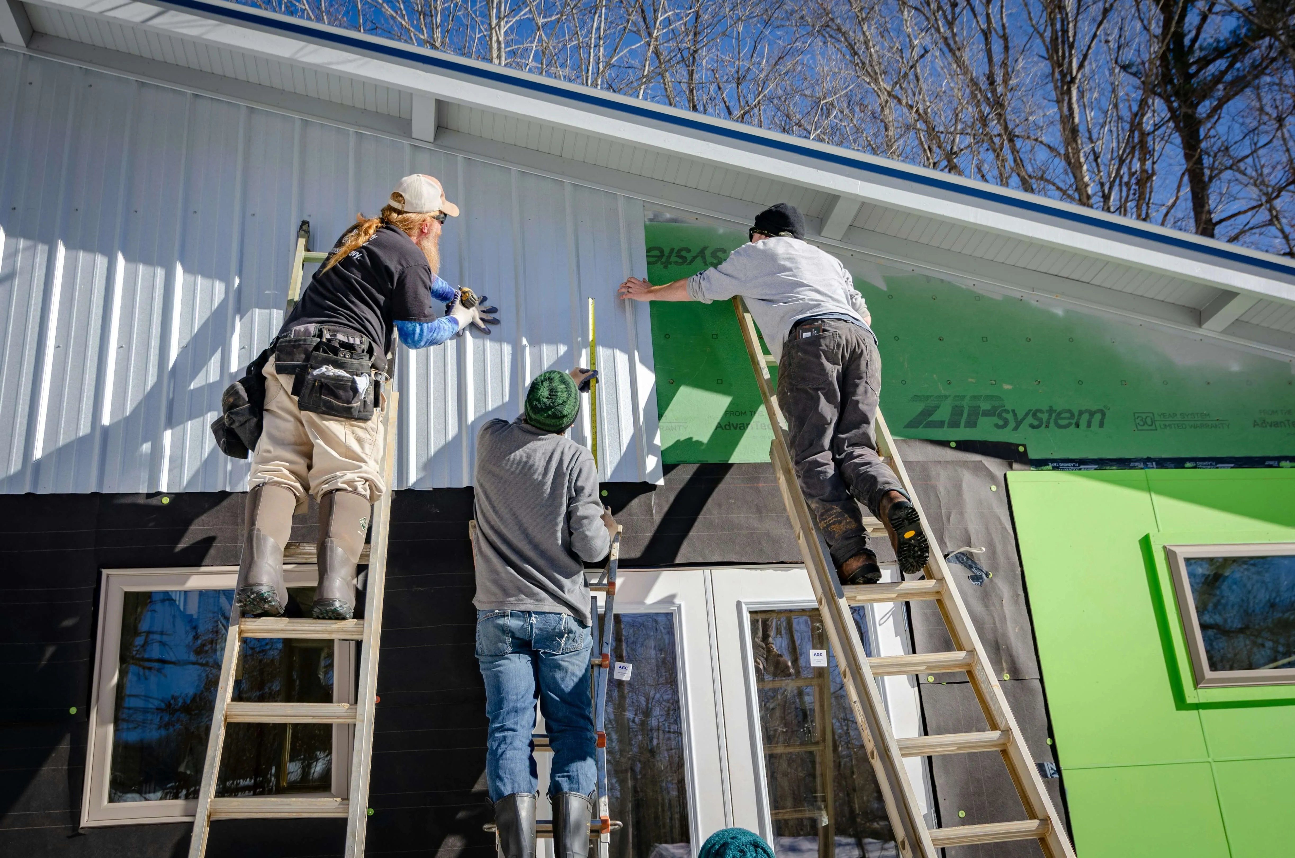 Three workers on ladders installing white siding siding on a house under construction.