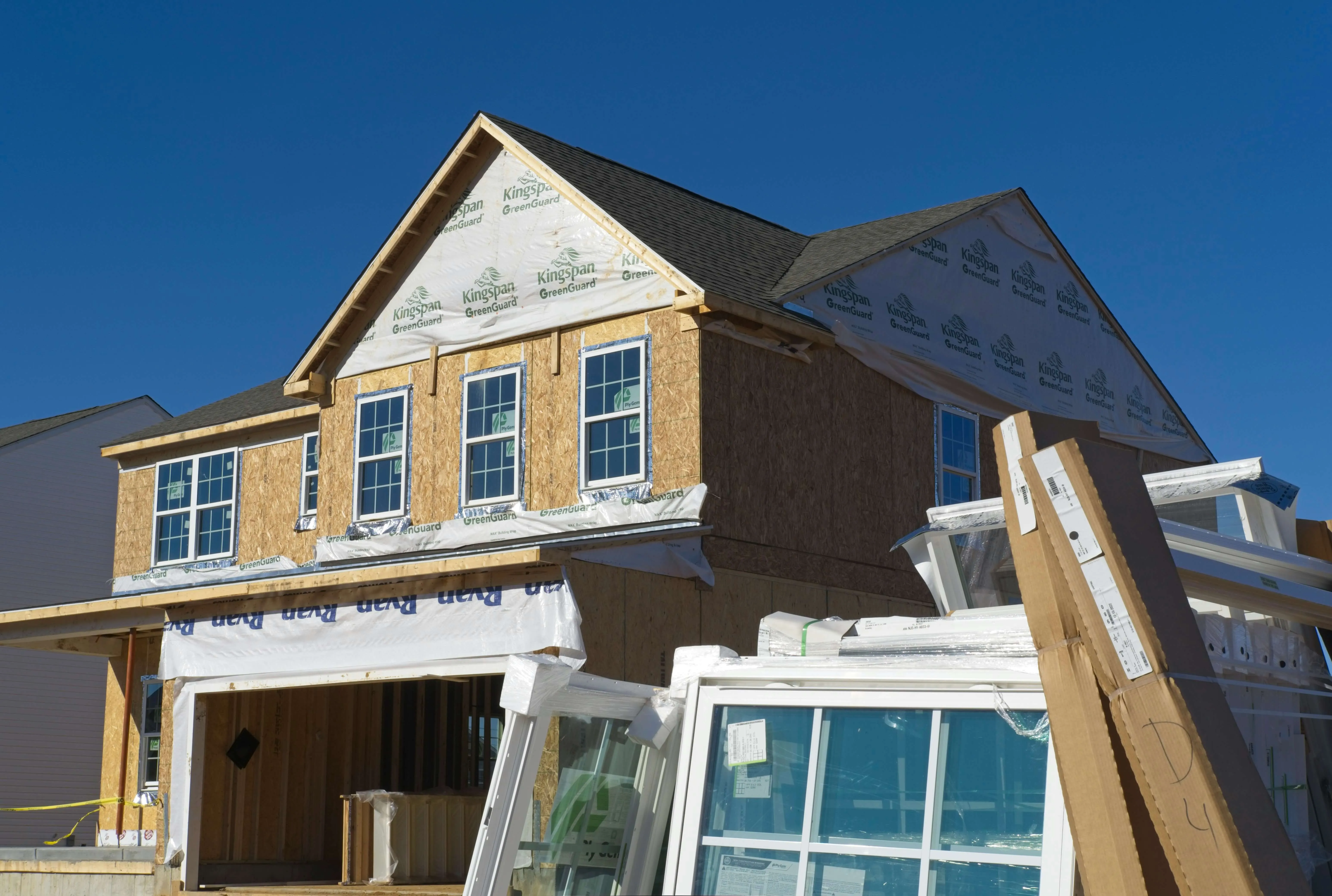 Two-story house under construction with wood framing, installed windows, and building materials outside.