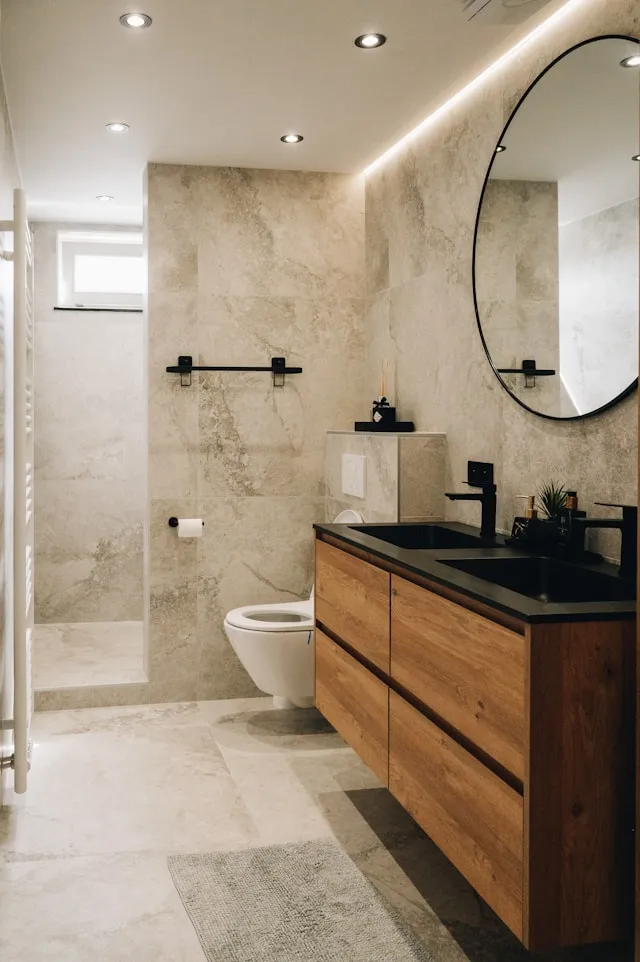 Modern bathroom with beige tiles, wooden vanity, black sinks, round mirror, and walk-in shower.