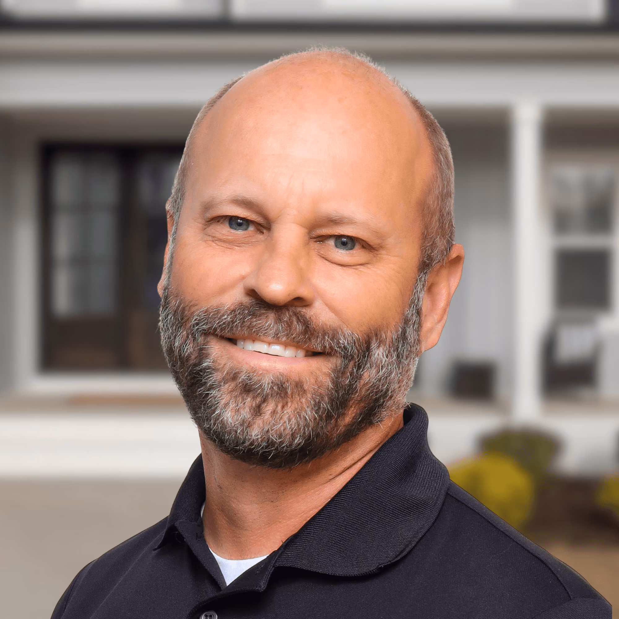 Smiling middle-aged man with beard and blue eyes wearing a black polo shirt against blurred outdoor background.