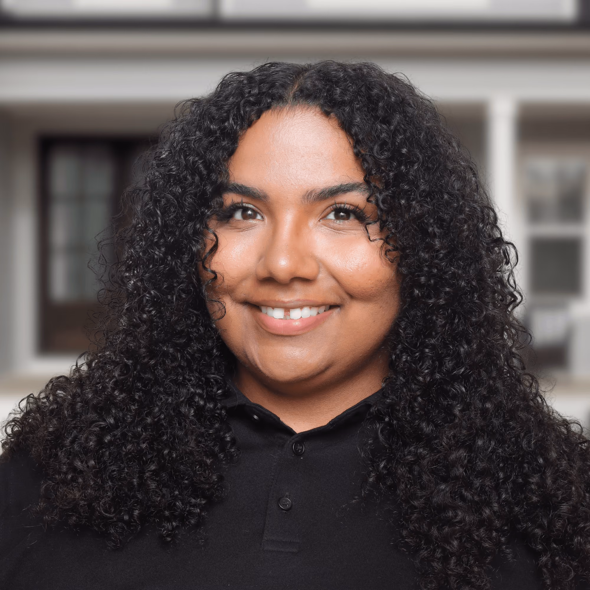 Smiling woman with long curly hair wearing a black collared shirt in front of a blurred house.