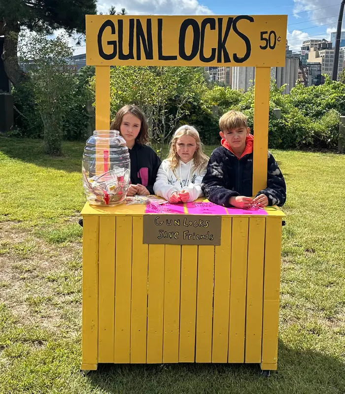 Three children standing behind a yellow wooden stand modeled after a lemonade stand, with signs that read "Gunlocks 50¢" and "Gunlocks Save Friends."
