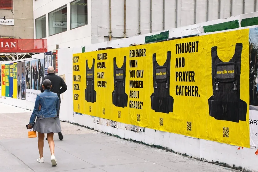 Pedestrians walk past a wall covered with yellow posters depicting black vests labeled 'NEW LIFE VEST' alongside text questions about keys, wallet, vest, business casual, and grades.