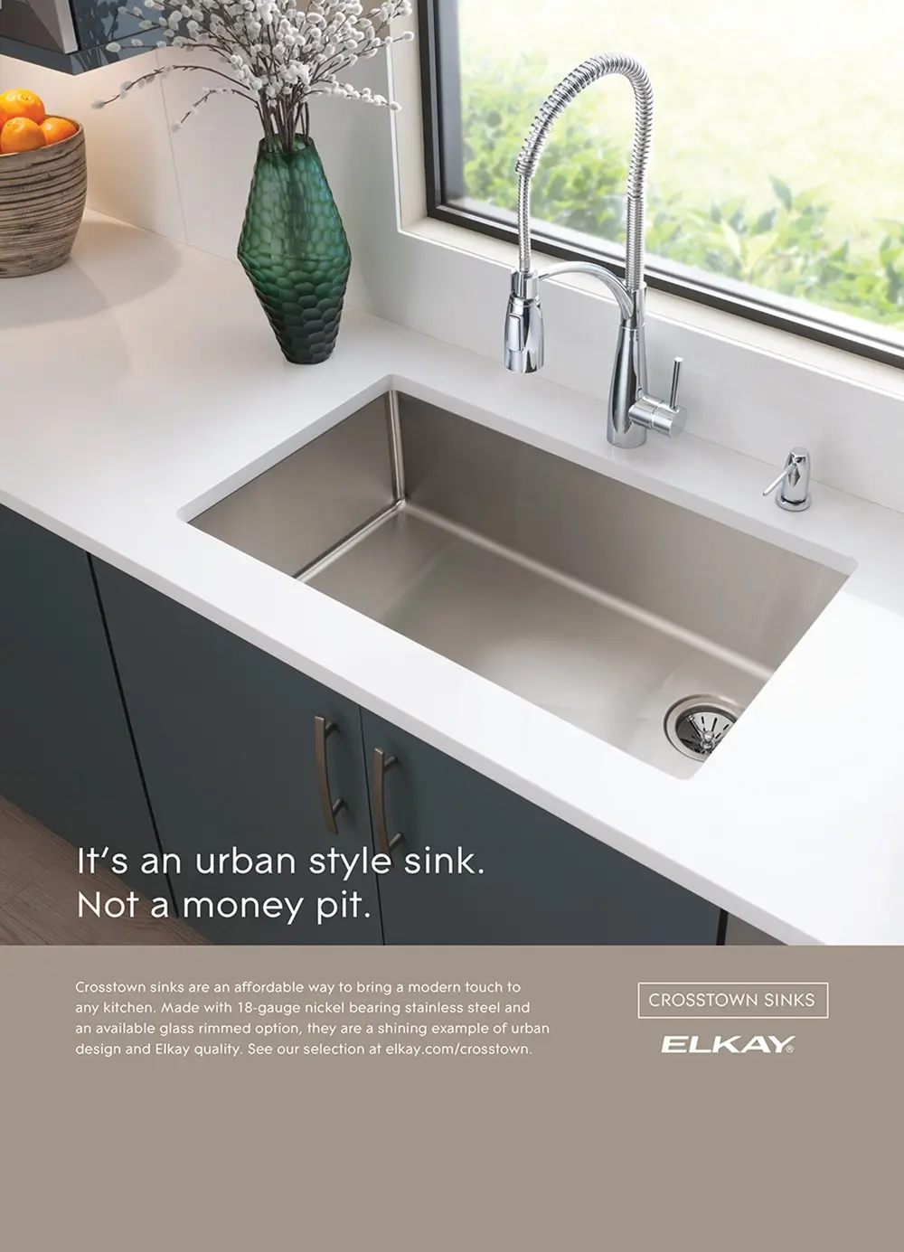 Modern stainless steel kitchen sink with a spring coil faucet, green textured vase with white flowers, and a bowl of oranges on a white countertop by a window.