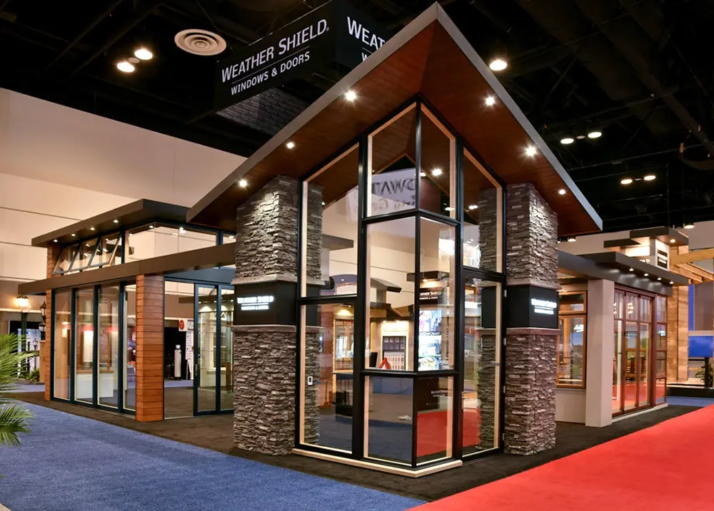 Modern exhibition booth for Weather Shield featuring large windows, stone pillars, wood paneling, and angled roof under indoor lighting.