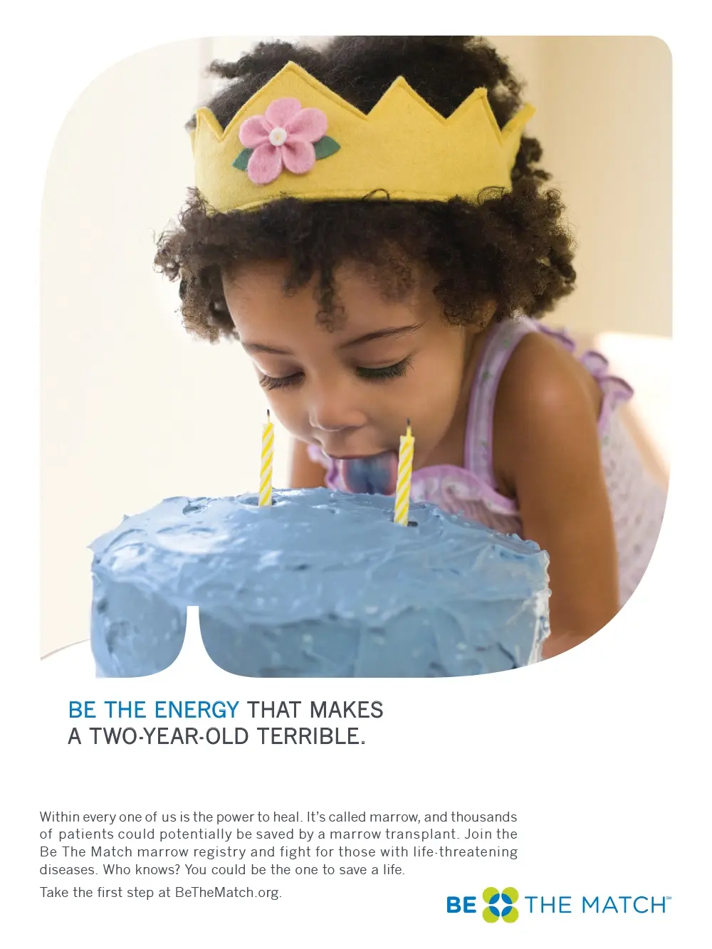A young child wearing a yellow crown with a pink flower leans forward to lick the blue frosting off a birthday cake with two lit candles.