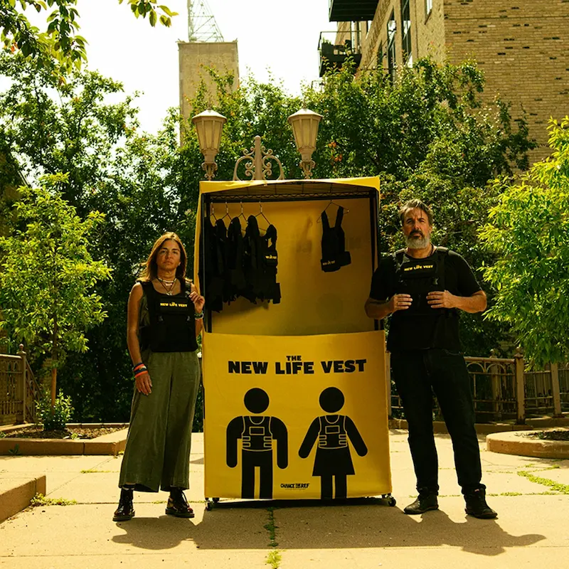 Two people wearing black life vests stand beside a yellow kiosk labeled 'The New Life Vest' with life vests hanging inside.