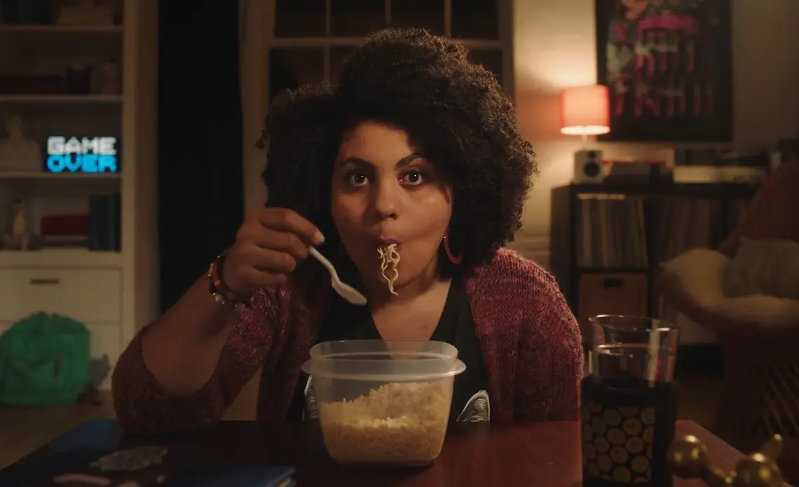 Woman with curly hair eating noodles from a plastic container at a table in a cozy room.