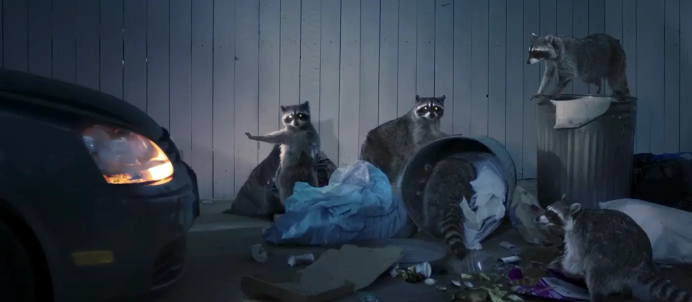 Five raccoons scavenging around trash bags, overturned trash cans, and scattered waste near a car with its headlights on.