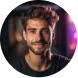 Smiling man with short, styled hair and a beard, wearing a dark shirt in a dimly lit setting with colorful blurred lights in the background.