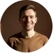Smiling man with short hair and beard wearing a brown sweater against a dark background.