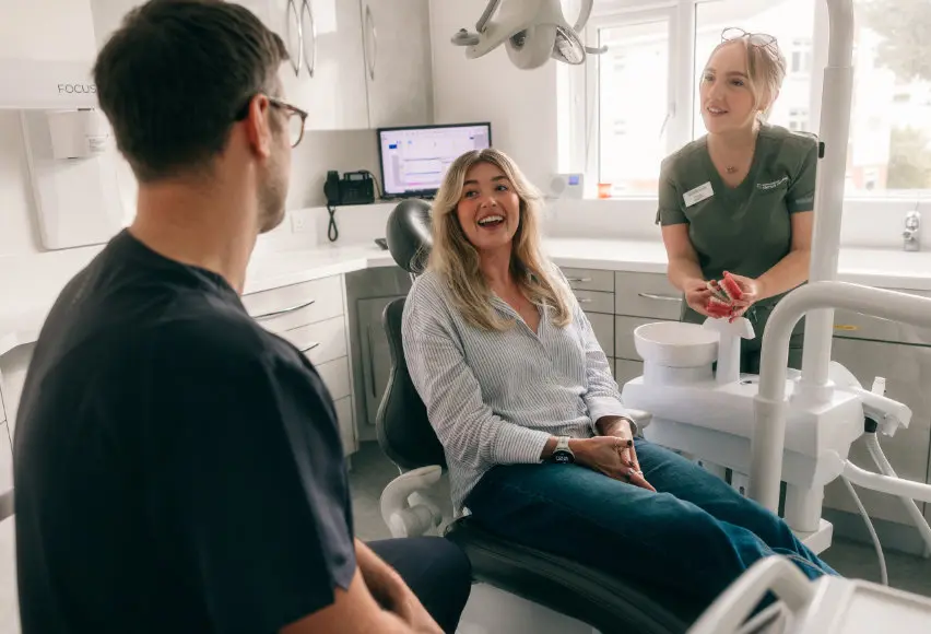 Woman smiling in a dental chair talking to two dental professionals in a bright clinic.