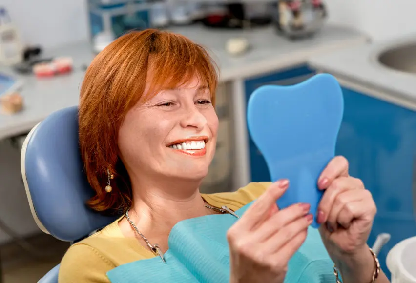 Smiling woman with red hair sitting in dental chair holding a blue tooth-shaped mirror.