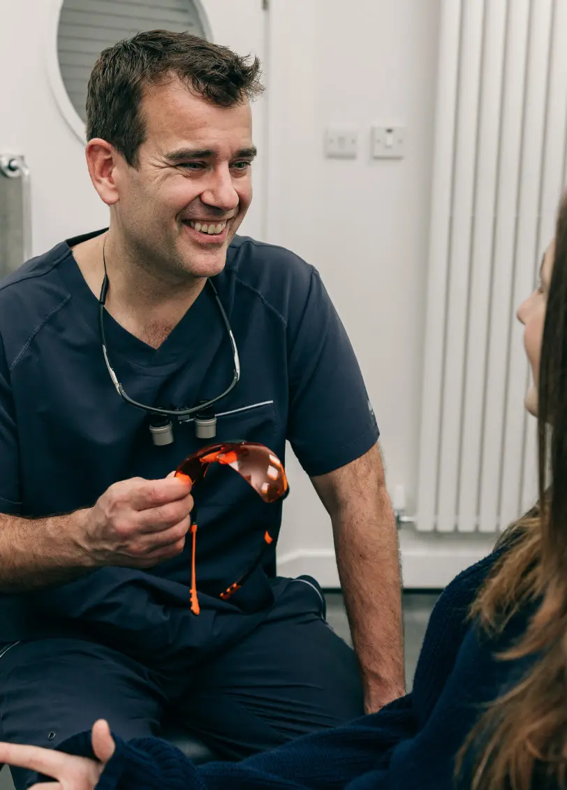 Smiling male dentist in navy scrubs holding orange protective glasses talking to a patient.