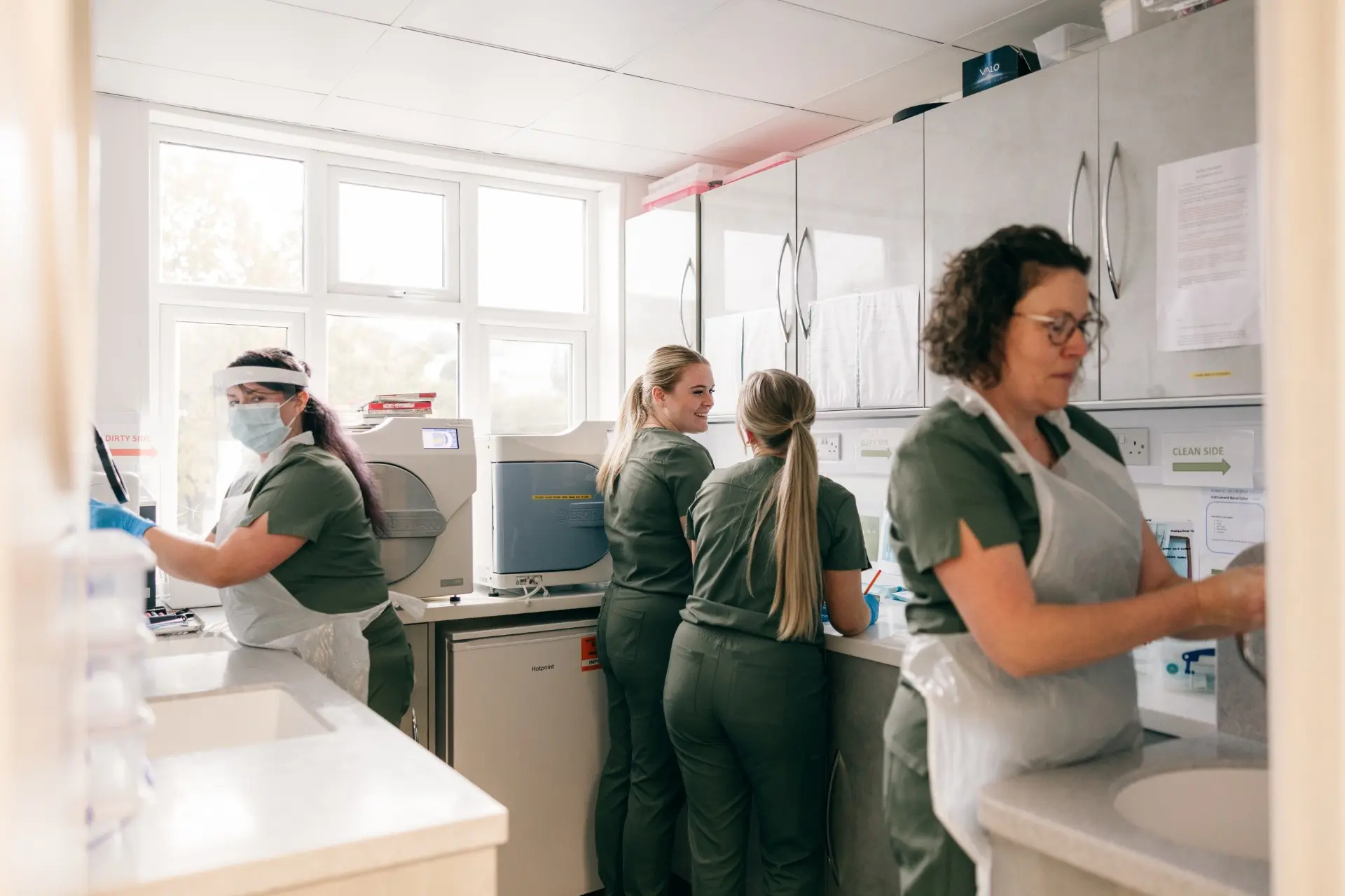 Four healthcare workers in green scrubs and aprons working in a sterile clinical lab with equipment.