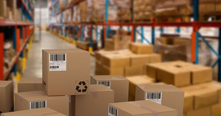 Stacks of cardboard boxes with barcodes in a warehouse, complemented by neatly organized shelves full of more boxes in the background. The scene is well-lit, with the front boxes in sharp focus. Freight paperwork is seen clipped to some of the boxes, while the rest of the warehouse remains slightly blurred.