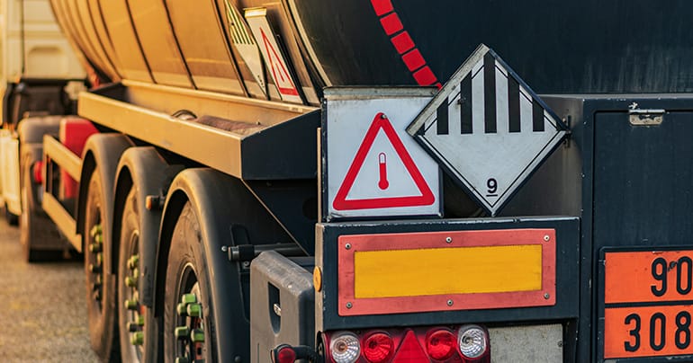 A close-up view of the rear side of a black tanker truck. The truck displays various safety signs including a thermometer warning, a black and white striped diamond, and an orange plate with numbers. Freight paperwork is secured nearby. The rear lights are visible, and sunlight casts shadows on the truck.