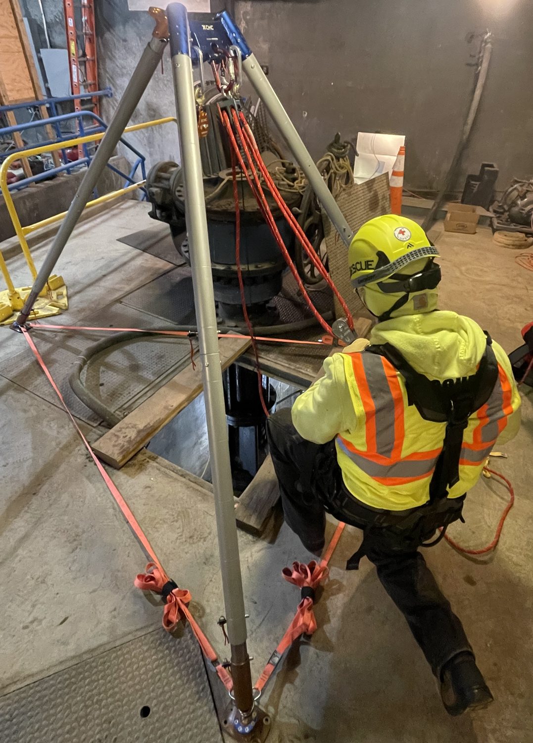 Rescue worker in high-visibility gear kneels near tripod rigging above a confined space opening.