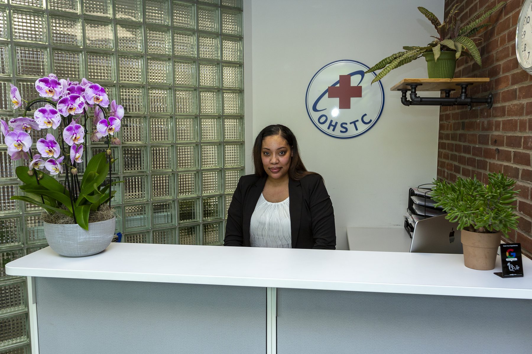 Receptionist seated behind a white desk with orchid and succulent plants in a modern office lobby.