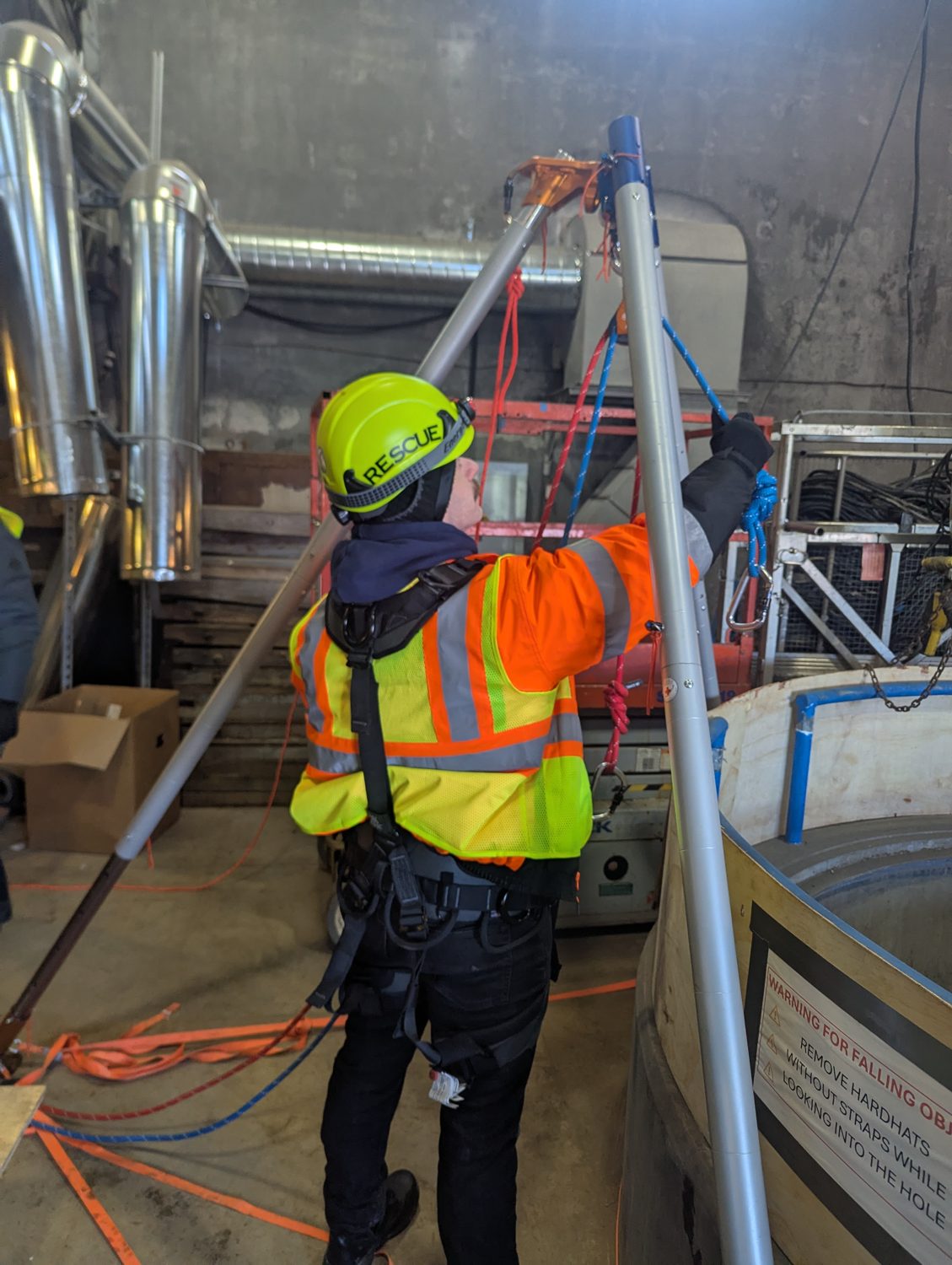 Worker in rescue helmet and safety vest handling ropes on metal tripod over industrial hole.