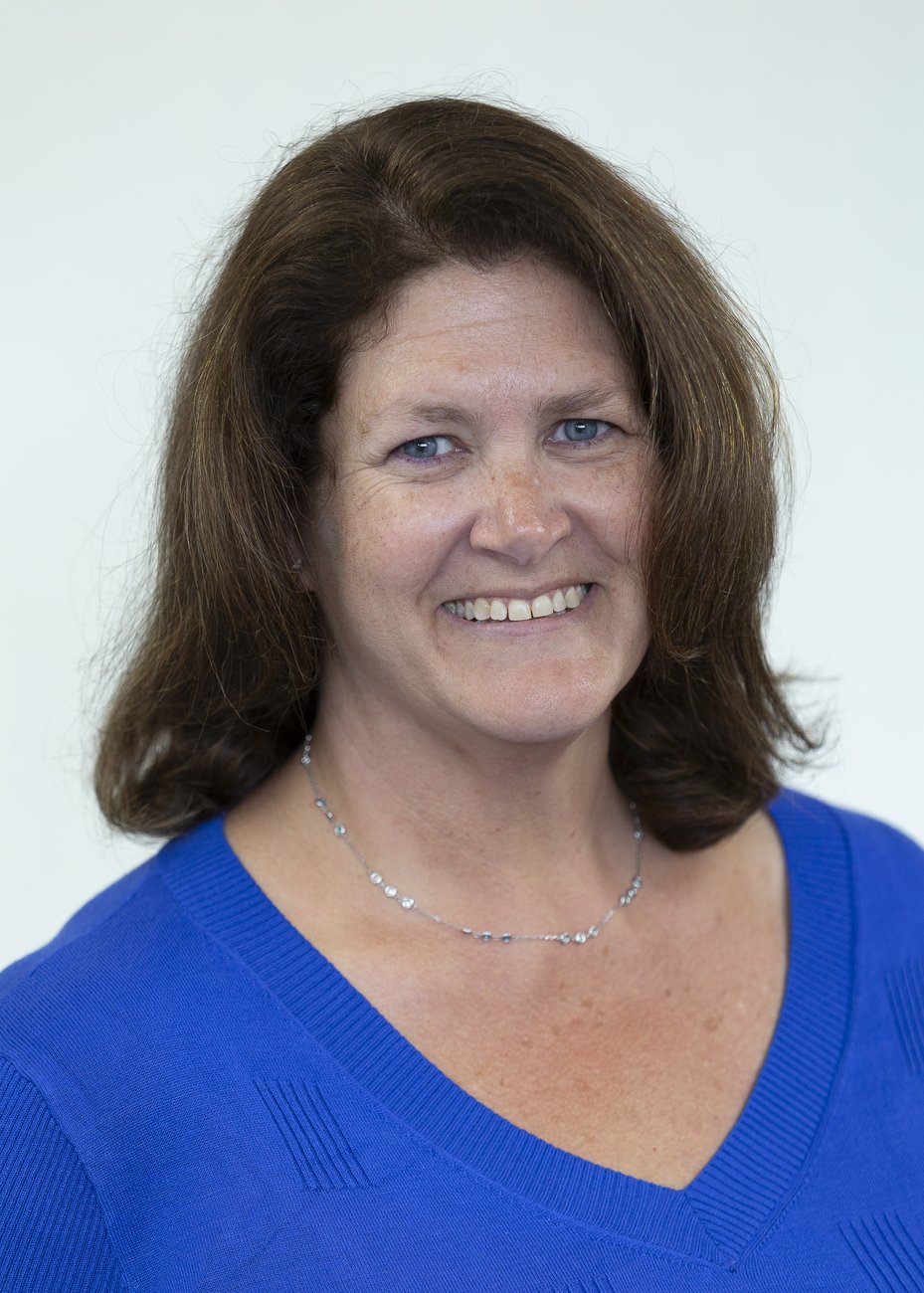Smiling woman with brown hair wearing a blue top and a delicate silver necklace on a white background.