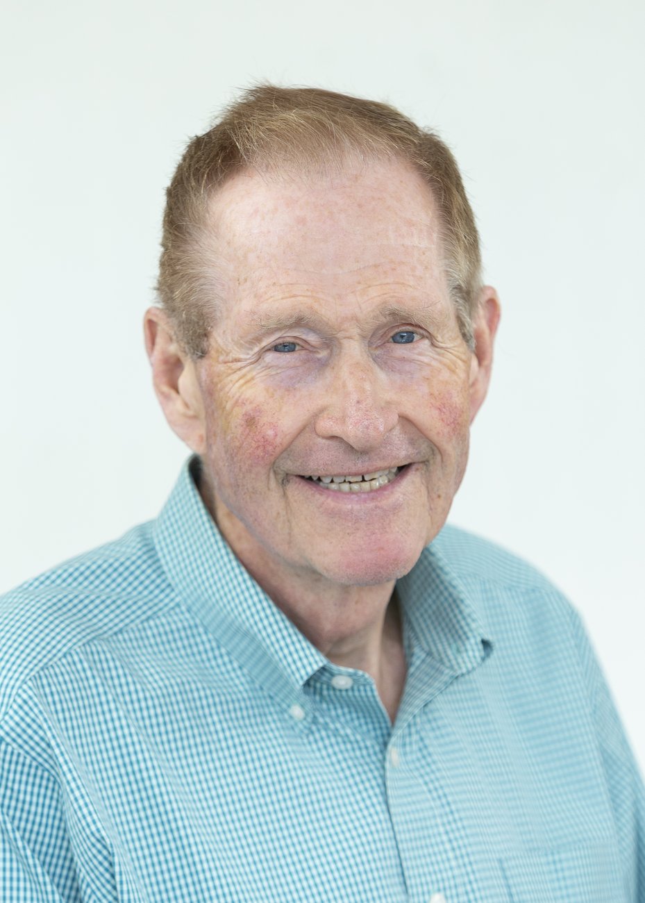 Smiling elderly man with light blue eyes wearing a blue checkered shirt against a plain background.