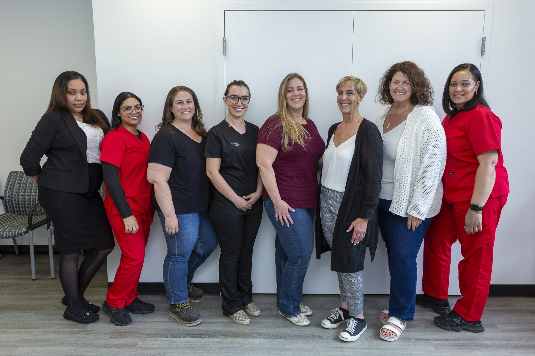Eight women smiling and standing side by side indoors against a white background.