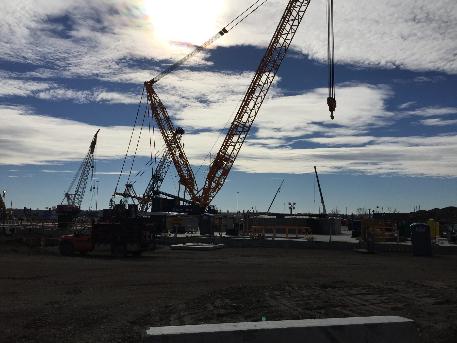 Multiple construction cranes at an outdoor job site under a partly cloudy sky with the sun shining.